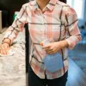 A woman in a plaid shirt cleaning a kitchen countertop with a cloth and holding a spray bottle, representing the rising demand for home care maids in Singapore.