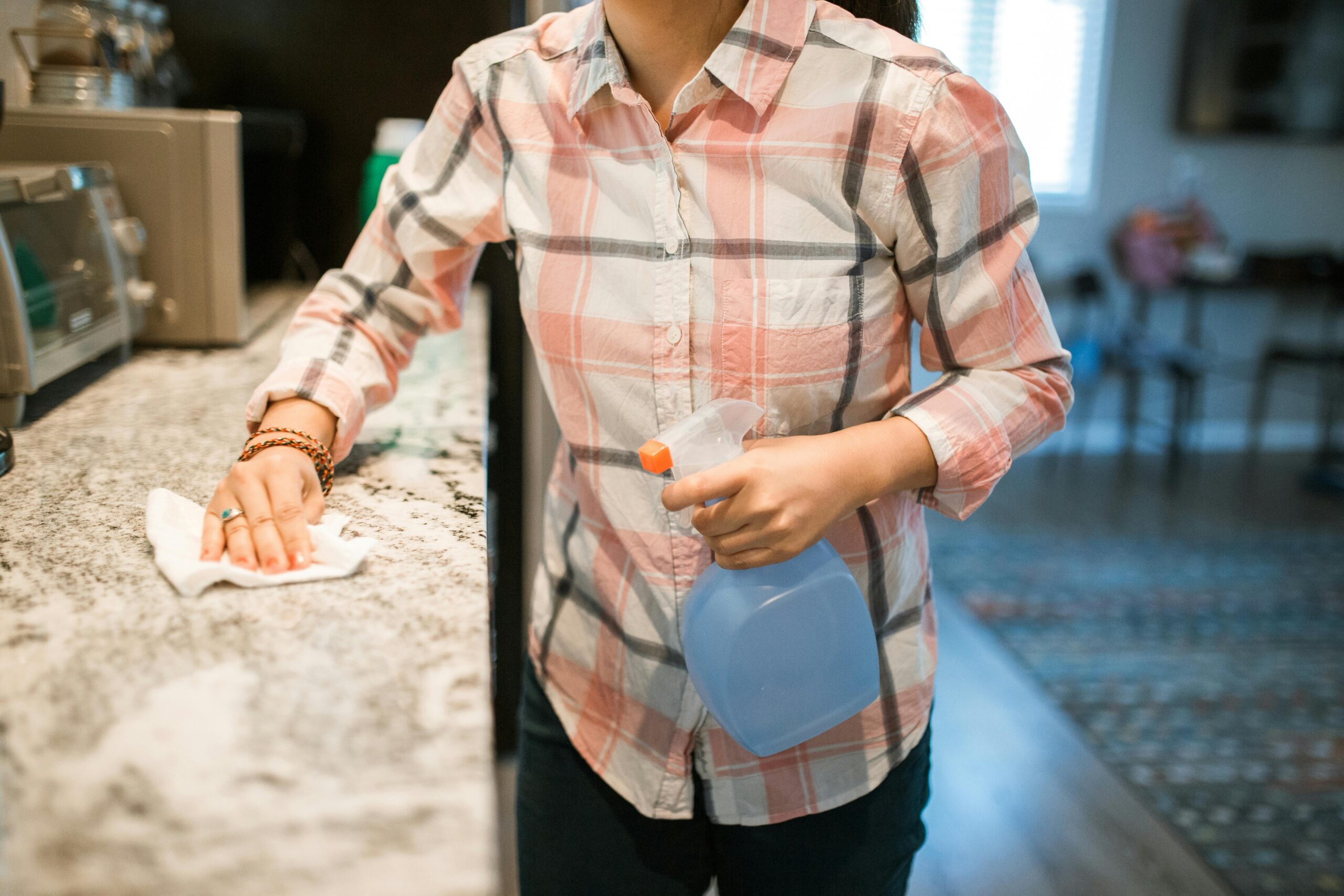 A woman in a plaid shirt cleaning a kitchen countertop with a cloth and holding a spray bottle, representing the rising demand for home care maids in Singapore.